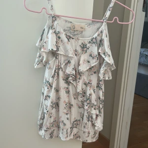 Supersöt blommig volangtop - Supersöt blommig topp från Ü i vitt med rosa och gröna blommor. Toppen har smala axelband, volangdetaljer framtill, banden är inte justerbara🙌 Tröjan är använd några få gånger men det är nästan inga täcken på användning, pris kan diskuteras
