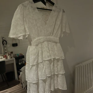 Vit volangklänning med fint mönster  - Superfin vit kortklänning med v-ringning, volanger och spetsmönster. Klänningen har korta ärmar och markerad midja, perfekt till sommaren