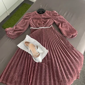 Glittrig vinröd långklänning med puffärm - Superfin vinröd långklänning med glittrigt tyg och plisserad kjol. Klänningen har puffiga långärmar och ett snyggt bälte med pärlor i midjan. Perfekt för dig som vill sticka ut med en elegant och trendig look. Skorna är i storlek 34 dem är för 100kr om du vill få dem med dem är i nyskick