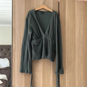 Grön omlottopp från Gina Tricot - Snygg grön omlottopp från Gina Tricot i storlek S. Toppen har lång ärm, v-ringad front och knytband i midjan.  Materialet är lätt och har en diskret struktur. 
