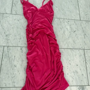 Rosa långklänning med rynkade detaljer - Säljer en snygg rosa långklänning med smala axelband och v-ringad hals. Klänningen har rynkade detaljer längs sidorna som ger en cool och trendig look. Materialet känns mjukt och stretchigt, perfekt för dig som vill sticka ut med färg och form.