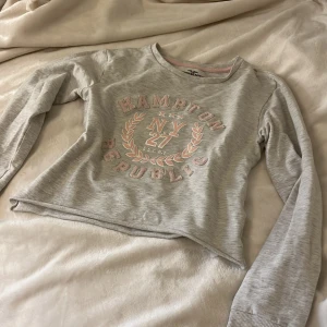 Grå lite kortare tjocktröja - Ljusgrå tröja från Hampton Republic med rosa och vita detaljer och broderad text framtill. Tröjan har rund halsringning och är avcroppad nertill vilket ger den en lite kortare och snyggare look.