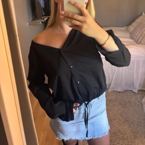 Svart blus med knytning  - Svart långärmad blus med v-ringning och knappar framtill från The Black Label Collection av lager 157. Blusen har en dragsko i midjan för en snygg passform och kan bäras offshoulder för en trendig look. Perfekt till jeans eller kjol.