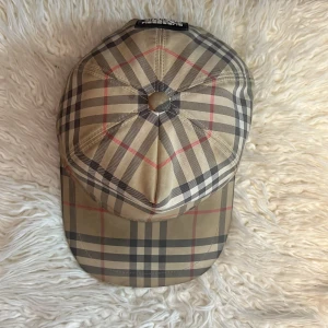 Burberry rutig keps i beige - Snygg keps från Burberry med klassiskt rutigt mönster i beige, svart och rött. Justerbar baktill med svart spänne och tydlig Burberry London England-patch. Perfekt accessoar för en trendig look.