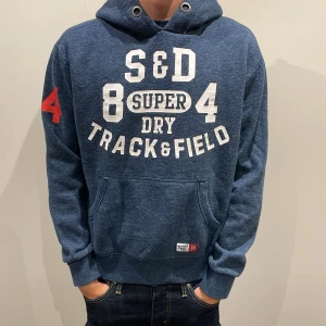 Blå Superdry hoodie med tryck - Rikgtigt snygg blå hoodie från Superdry med vit text och tryck på bröstet samt röd siffra på ärmen. Nypris 900kr vårt pris 700kr. Bara att skriva ifall du har några frågor!