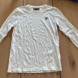 Vit långärmad tröja från Lyle & Scott - Vit långärmad tröja från Lyle & Scott med klassisk rund hals och den ikoniska gulsvarta logon broderad på bröstet. Tröjan är gjord i mjuk bomull och har en clean, enkel look som funkar till jeans eller shorts. Perfekt för dig som gillar stilrena plagg.