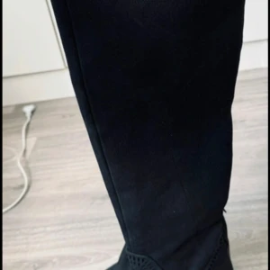 Svarta höga mockastövlar - Snygga svarta stövlar i mocka med högt skaft som går över knäet. De har en låg klack och rund tå, samt diskreta sömdetaljer upptill. Perfekta till jeans eller kjol för en trendig look.