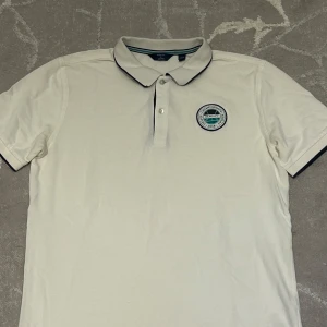 Vit pikétröja GANT x Båstad - Snygg vit pikétröja från GANT x Båstad med klassisk krage och två knappar. Tröjan har en broderad patch på bröstet och tunna svarta detaljer på krage och ärmslut. Perfekt för en sportig och clean look.