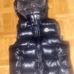 Svart dunväst från Moncler med huva - Säljer en svart, glansig dunväst från Moncler med huva och dragkedja framtill. Västen har två fickor med dragkedja och ikonisk Moncler-logga på bröstet. Insidan har en cool serietidningsdetalj. Perfekt för dig som vill ha en snygg och varm look.