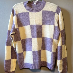 Lila och vit rutig stickad tröja Lindex - Supermysig stickad tröja från Lindex i lila och vitt med rutigt mönster. Tröjan har rund halsringning, ribbade muddar och lång ärm. Perfekt för dig som gillar en chill och trendig vibe.