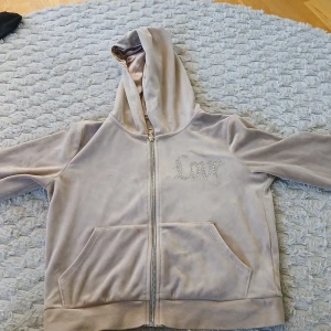 Beige hoodie med strass från Lindex - Säljer en beige hoodie från Lindex med mjuk, sammetsliknande yta och glansigt foder i huvan. Framsidan har strassdetaljer som bildar ordet 'Love'. Tröjan har dragkedja, huva och två fickor framtill. Perfekt för dig som gillar comfy och snygg stil. Finns lika dan fast i byxa