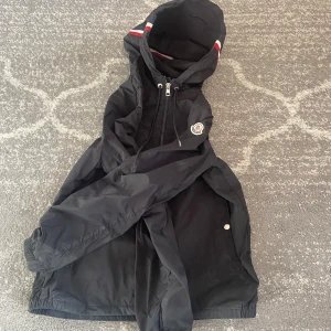 Moncler jacka - Svart vindjacka från Moncler med huva och dragsko, klassisk logga på ärmen och snygga detaljer vid fickorna. Jackan har dragkedja framtill och är perfekt för dig som vill ha en stilren och sportig look. Passar till både vår och höst.