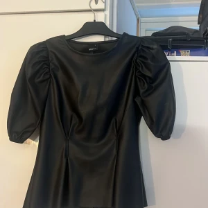 Svart puffärm blus från Gina Tricot - Snygg svart blus från Gina Tricot i storlek S med puffiga korta ärmar och rund hals. Tillverkad i skinnliknande syntetmaterial som ger en cool vibe. Figurnära passform och dragkedja i ryggen. Perfekt för dig som gillar statement-plagg med edge.