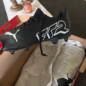 Puma Future 7 Play fotbollsskor svart/vit ( oanvända ) - Snygga Puma Future 7 Play fotbollsskor i svart med vita detaljer och logga. Skorna har snörning, mönstrad ovandel och fasta dobbar för spel på både gräs och konstgräs. Lätta och smidiga med modern design, perfekta för dig som vill sticka ut på planen. Helt oanvända och oprövade.