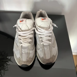 Nike Air Max dawn sneakers i vitt och grått - Nike Air Max dawn sneakers i vitt och ljusgrått med klassisk swoosh på sidan. Ovandel i textil och mocka, chunky sula med synlig Air Max-enhet och vita snören. Retroinspirerad design med orange Nike-logga på plösen. Perfekta för dig som gillar streetwear.