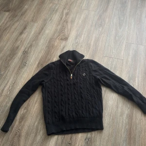 Svart stickad half zip tröja Morris - Svart stickad tröja från Morris med kabelstickat mönster och half zip i halsen. Tröjan har ribbade muddar och hög krage, samt en liten logga på bröstet. Perfekt för dig som gillar klassisk stil med en modern twist.