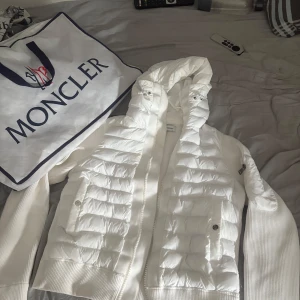 Vit Moncler pufferjacka med huva - Säljer en clean vit pufferjacka från Moncler med stickade ärmar och vadderad framsida. Jackan har dragkedja, stor huva och fickor med tryckknapp. Moncler-logga på ärmen och cool detaljficka på armen. Perfekt för dig som vill ha en stilren och exklusiv look. Storlek M men passar som S.