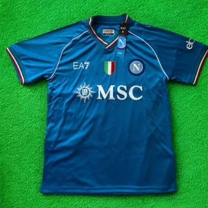 Napoli matchtröja EA7 blå - Napoli fotbollströja från EA7 i blått med vita detaljer och klubbmärke på bröstet. Tröjan har korta ärmar, V-ringad krage med italienska färger och tryckta sponsorer. Tillverkad i lätt och ventilerande material, perfekt för match eller träning.