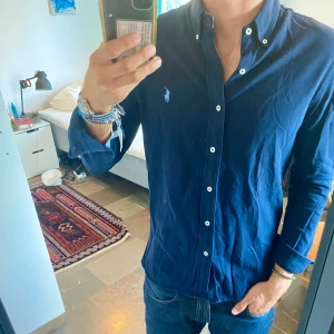 Mörkblå skjorta från Ralph Lauren - Säljer en klassisk mörkblå skjorta från Ralph Lauren med button-down krage och vita knappar. Skjortan har broderad logga på bröstet och är tillverkad i ett lätt bomullsmaterial. Perfekt för dig som gillar stilrena och tidlösa plagg.