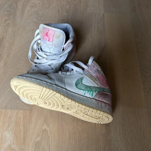 Nike Air Jordan 1 med färgstänk - Unika Nike Air Jordan 1 sneakers med coola färgstänk i grönt och rosa, samt detaljer i beige och gult. Skorna har snörning, hög siluett och en mix av skinn och textil. Swoosh-loggan är grön med drip-effekt och Air Jordan-loggan syns i rosa på sidan.