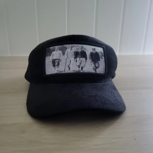 Svart keps Emiliano Padenza vintageprint - Svart meshkeps från Emiliano Padenza med vintageinspirerat svartvitt foto framtill. Kepsen har böjd skärm, justerbar baksida och ventilerande meshpaneler. Perfekt för dig som gillar retrostil och unika detaljer.