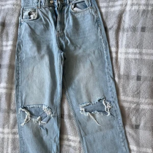 Ljusblå ripped jeans från Gina Tricot - Säljer ett par ljusblå jeans från Gina Tricot med stora slitningar och fransade hål på båda benen. Klassisk femficksmodell med raka ben och hög midja. Perfekta för en avslappnad och trendig look.