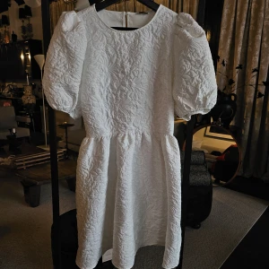 Vit quiltad kortklänning med puffärm - Superfin vit kortklänning med quiltad struktur och broderade blommor. Klänningen har puffiga korta ärmar, rund halsringning och knäppning bak. Den har en markerad midja och voluminös kjol som ger en söt och trendig look.