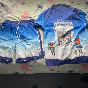 Casablanca blå kortärmad skjorta - Snygg kortärmad skjorta från Casablanca med blå och vit gradient, tryckta snäckskal och coolt Paris-motiv på ryggen. Skjortan har knappar framtill och matchar perfekt med shorts i samma stil. Perfekt för sommaren och chill vibes.