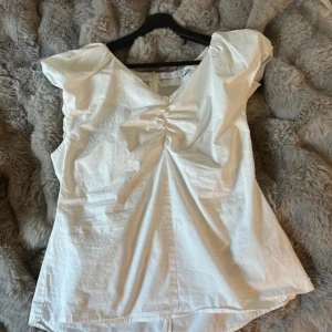 Vit blus från Coster Copenhagen - Supersöt vit blus från Coster Copenhagen med v-ringning, puffiga axlar och snygg rynkdetalj framtill. Blusen har korta ärmar och dragkedja i sidan. 