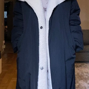 Svart lång pufferjacka med teddykrage - Lång svart pufferjacka med oversized passform och stor vit teddykrage. Jackan har knappar framtill och är helfodrad med mjukt teddyfoder längs insidan och kragen. Perfekt för kalla dagar när du vill vara både varm och stilren.