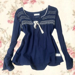 Mörkblå blus med broderi och knyt - Supersöt mörkblå blus från Odd Molly med vita broderier över bröstet och en dekorativ knytning framtill. Långärmad modell med avslappnad passform och rundad halsringning. Perfekt för dig som gillar boho-stil och unika detaljer.