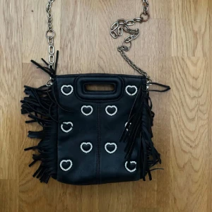 Svart axelväska med pärlhjärtan och fransar - Svart axelväska från MAJE med silverfärgad kedja, pärlformade hjärtan på framsidan och långa fransar på sidorna. Väskan har ett litet handtag upptill och unik design som sticker ut.