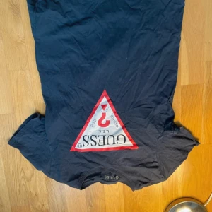 Svart t-shirt från Guess med logga - Svart t-shirt från Guess i slim fit med klassisk triangel-logga framtill i rött och vitt. T-shirten är kortärmad och tillverkad i mjuk bomull, perfekt för en clean och stilren look. Snygg passform och ikonisk design.