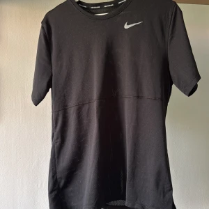 Svart Nike t-shirt för löpning - Svart t-shirt från Nike med klassisk Swoosh-logga på bröstet. Tillverkad i lätt och ventilerande polyester, perfekt för löpning eller träning. Kortärmad modell med rund hals och diskret meshstruktur för extra andningsförmåga.