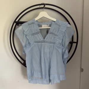 Ljusblå volangblus från Neo Noir med prislapp - Supersöt ljusblå blus från Neo Noir med volangdetaljer och v-ringning. Blusen har korta ärmar och är tillverkad i ett tunt, luftigt material som passar perfekt till varmare dagar. Helt ny med prislappen kvar