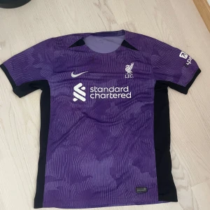 Liverpool tredjetröja 23/24 - Liverpool FC matchtröja i lila med svarta detaljer. Tröjan har korta ärmar, vit logga och sponsortryck på bröstet samt ett subtilt mönster över hela tyget. Tillverkad i lätt och ventilerande material, perfekt för träning eller match. Passar både S och M. Pris kan diskuteras