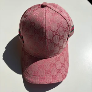 Rosa keps från Gucci med GG-mönster - Snygg rosa keps från Gucci med ikoniskt GG-monogram över hela kepsen. Klassisk böjd skärm och justerbar rem baktill. Sidorna har gröna och röda ränder som ger den extra Gucci-vibe. Tillverkad i bomull och polyester för en lyxig känsla.