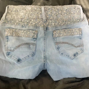 Jeansshorts  - Unika ljusblå jeansshorts från West M&M  med snygga spetsdetaljer och broderier i midjan och på bakfickorna. Shortsen har råa kanter nertill och glittrande stenar på bakfickorna för extra bling. Perfekta för dig som vill sticka ut i sommar!