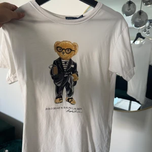Vit Polo Bear t-shirt från Ralph Lauren - Vit t-shirt från Ralph Lauren med ikoniskt Polo Bear-tryck på bröstet. Klassisk rund hals och korta ärmar. T-shirten är i mjuk bomull och har en avslappnad passform. Perfekt för dig som gillar stilrena plagg med en twist.
