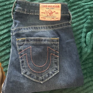 True Religion blå jeans med kontrastsöm - Säljer ett par klassiska blå jeans från True Religion med ikonisk kontrastsöm i rött på bakfickan och stor patch bak i midjan. Jeansen har en snygg passform och är tillverkade i mjukt denimtyg. Perfekta för dig som gillar streetwear och vill sticka ut. 