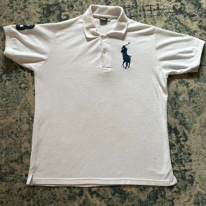 Vit Polo Ralph Lauren t-shirt - Vit t-shirt från Polo Ralph Lauren med klassisk polokrage och två knappar. Stor broderad marinblå polospelare på bröstet och en broderad siffra 3 på ena ärmen. Tillverkad i bomull med normal passform, perfekt för en clean och sportig look.