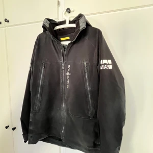 Svart vindjacka från Sail Racing - Svart vindjacka från Sail Racing med hög krage och huva. Jackan har flera dragkedjeförsedda fickor framtill och logga på bröstet samt ärmen. Snygg och sportig look, perfekt för blåsiga dagar. Materialet känns slitstarkt och jackan har en clean design.