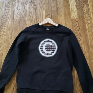 Colmar sweatshirt  - Svart sweatshirt från Colmar Originals med rund hals och lång ärm. På bröstet finns ett stort cirkulärt tryck med texten 'Colmar Originals' i vitt och grått. Tröjan har ribbade muddar och är gjord  bomull, perfekt för chill dagar.