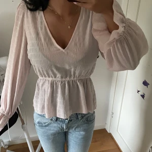 Ljusrosa blus med v-ringning - Supersöt ljusrosa blus med v-ringad hals och voluminösa långärmar. Smockad överdel och lätt rynkad midja ger en snygg siluett. Perfekt att matcha med jeans för en chill look. Materialet känns lätt och luftigt.