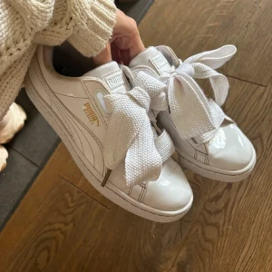 Sneakers  - Snygga vita sneakers från Puma med breda, platta snören och gulddetaljer på sidan. Skorna har en klassisk rund tå och är tillverkade i blankt syntetmaterial som ger en fräsch look. Perfekta för dig som vill sticka ut med coola detaljer.