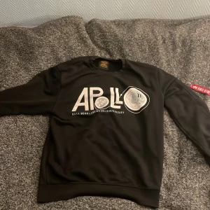 Svart Apollo sweatshirt Alpha Industries - Cool svart sweatshirt från Alpha Industries med stort Apollo-tryck på bröstet och NASA Moon Landing 50th Anniversary text. Unik detalj med röd 'Remove Before Flight'-tag på ärmen och dragkedja. Perfekt för dig som gillar rymdtema och streetwear.