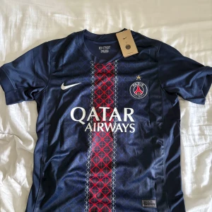 PSG Hemma Ställ 25/26 (#14 D. Doué) - Snygg Paris Saint-Germain fotbollströja med D. Doué och nummer 14 på ryggen. Marinblå med rött och vitt geometriskt mönster längs mitten, PSG-logga och Nike Swoosh på bröstet. Tillverkad i lätt och ventilerande polyester, perfekt för träning eller match.