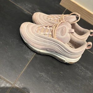 Nike Air Max 97 rosa sneakers strl 36 - Nike Air Max 97 sneakers i en ljusrosa färg med vågiga detaljer och synlig Air-sula. Skorna har snörning och en platt sula med svart yttersula. Materialet är syntet och textil, och modellen har en sportig vibe med ikonisk design. Perfekt för dig som gillar streetstyle och snygga sneakers.