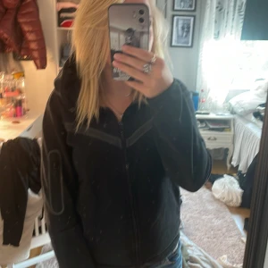 Svart Nike vindjacka med huva - Svart vindjacka från Nike med dragkedja framtill och huva. Jackan har en clean look med diskreta detaljer och är perfekt för dig som gillar sportig stil. Passformen är normal och materialet känns lätt och smidigt. Snygg att slänga på sig till jeans eller shorts.