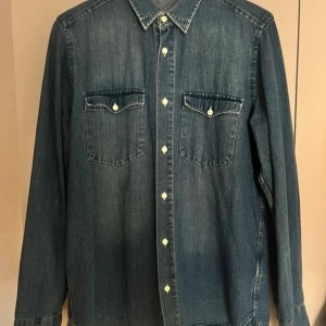 Jeansskjorta  - Snygg jeansskjorta från These Glory Days i klassisk blå denim. Skjortan har två bröstfickor med lock och knappar, samt vita knappar längs hela framsidan. Materialet är 100% bomull. Knappt använd!
